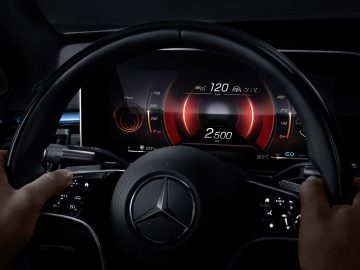 Close-up van de handen van een bestuurder op het stuur van een Mercedes-Benz S-Klasse, met de focus op het verlichte digitale dashboard met verschillende autogegevens.