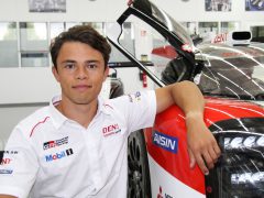 Jonge mannelijke autocoureur Nyck de Vries in een wit race-uniform, leunend op een rode en zwarte raceauto in een garage.