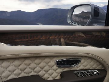 Binnenaanzicht van het deurpaneel van een Bentley Bentayga met gewatteerd leer en raambediening, met uitzicht op een bergachtig landschap via de zijspiegel.