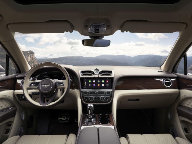 Luxe Bentley Bentayga-interieur met houten bekleding, lederen stoelen en modern dashboard tegen een bergachtige achtergrond.