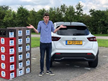 Man haalt zijn schouders op naast een stapel elektronica in een doos en een witte Nissan Juke op een parkeerplaats.