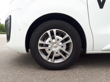Close-up van de linker voorband en lichtmetalen velg van een witte Opel Vivaro, geparkeerd op een asfaltoppervlak.
