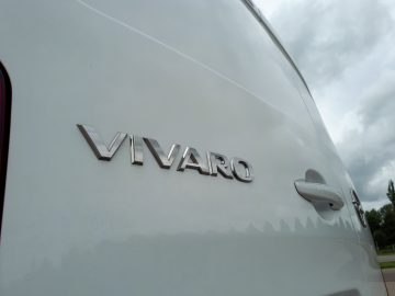 Close-up van het 'Opel Vivaro'-embleem op het witte zijpaneel van een voertuig, met een bewolkte lucht weerspiegeld op de achtergrond.