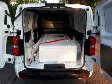 Een grote witte deur horizontaal geladen in de bagageruimte van een open witte Opel Vivaro-bestelwagen, beveiligd met een rode riem, bij daglicht.