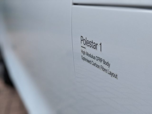 Close-up van een wit autopaneel met de tekst "Polestar 1 - cfrp-carrosserie met hoge modulus, geoptimaliseerde koolstofvezelindeling".