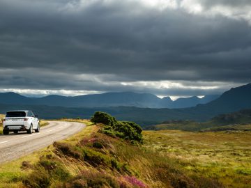 Een Range Rover rijdt over een bochtige weg door een bergachtig landschap onder een stormachtige hemel.