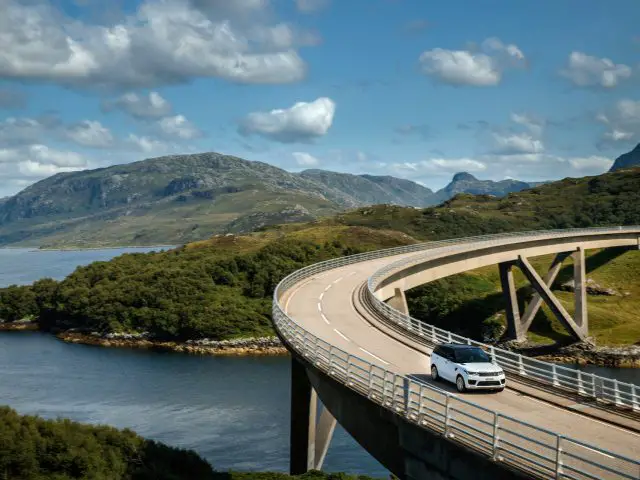Een Range Rover steekt een gebogen brug over met uitzicht op een meer met glooiende heuvels op de achtergrond onder een helderblauwe lucht.