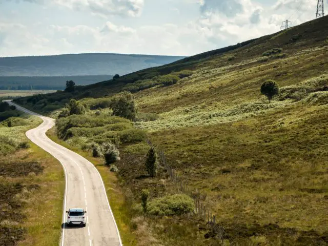 Een Range Rover rijdt over een bochtige weg door een heuvelachtig, groen landschap met schaarse bomen en verre windturbines.