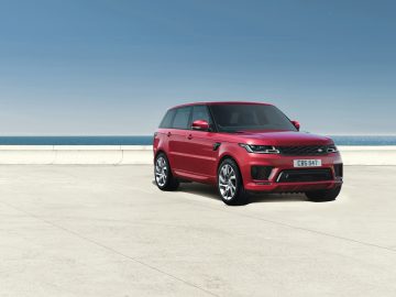 Red Range Rover geparkeerd op een betonnen ondergrond met een heldere lucht en een kalme zee op de achtergrond.