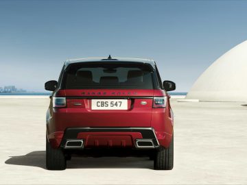 Een rode Range Rover staat geparkeerd voor een witte koepelstructuur, met op de verre achtergrond de skyline van de stad onder een helderblauwe lucht.