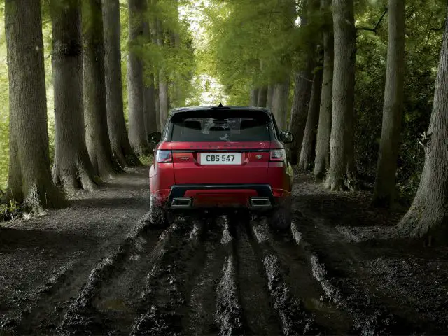 Een rode Range Rover geparkeerd op een modderig pad in een dicht bos, omgeven door hoge bomen, wat zijn robuuste buitencapaciteiten benadrukt.
