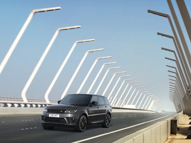 Een zwarte Range Rover die over een moderne brug met opvallende witte bogen rijdt onder een helderblauwe lucht.