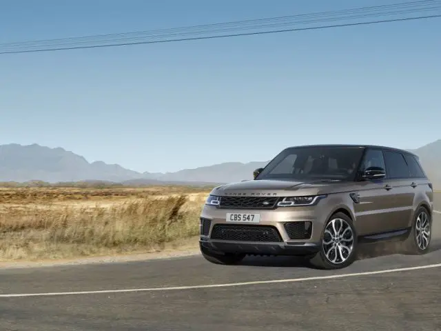 Een zilveren Range Rover die over een kronkelige weg rijdt, omringd door een dor landschap met bergen in de verte onder een helderblauwe lucht.