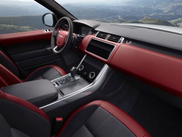 Binnenaanzicht van een Range Rover met een rood en zwart lederen dashboard, middenconsole en stuurwiel met een bergachtig landschap zichtbaar door de voorruit.
