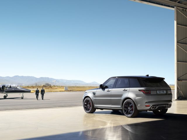 Een zilveren Range Rover geparkeerd buiten een hangar met een vliegtuig en twee mensen op de achtergrond, tegen een helderblauwe lucht en bergachtig terrein.