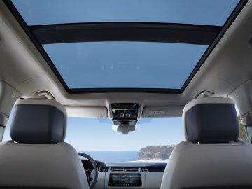 Binnenaanzicht van een Range Rover vanaf de achterbank met een open panoramisch schuifdak, dashboard en uitzicht op de oceaan door de voorruit.