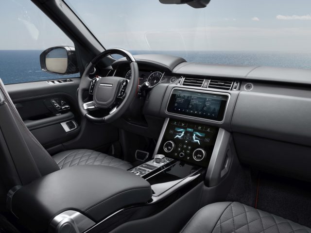 Binnenaanzicht van een Range Rover met het dashboard, het stuur en het multimediasysteem, met uitzicht op de oceaan buiten het raam.