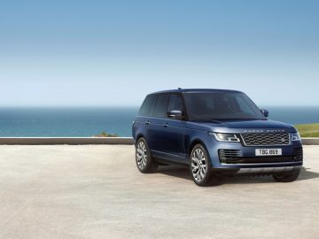 Een blauwe Range Rover geparkeerd op een kustweg met een helderblauwe lucht, de oceaan en een modern huis op de achtergrond.