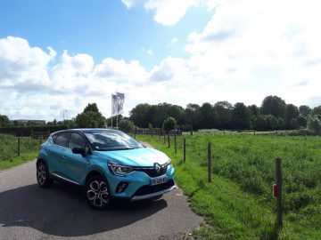 Een blauwgroen Renault Captur E-Tech geparkeerd op een landelijke weg met groene velden, een bewolkte lucht en een vlaggenmast op de achtergrond.