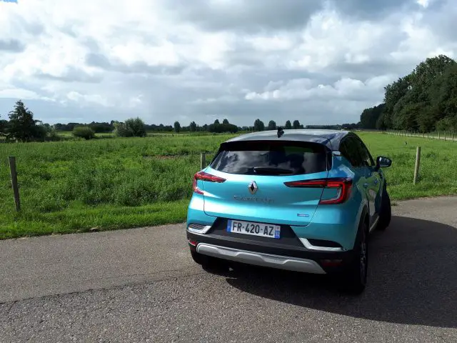 Een blauwe Renault Captur E-Tech geparkeerd op een weg naast een groen veld onder een bewolkte hemel.