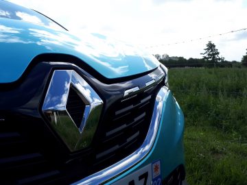 Close-up van de grille en het embleem van een blauwe Renault E-Tech-auto, met een wazig groen veld en bomen op de achtergrond.