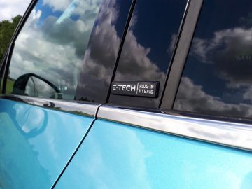 Close-up van de deur en het raam van een blauwe plug-in hybride auto, die wolken reflecteren, met een zichtbaar "E-Tech"-embleem.
