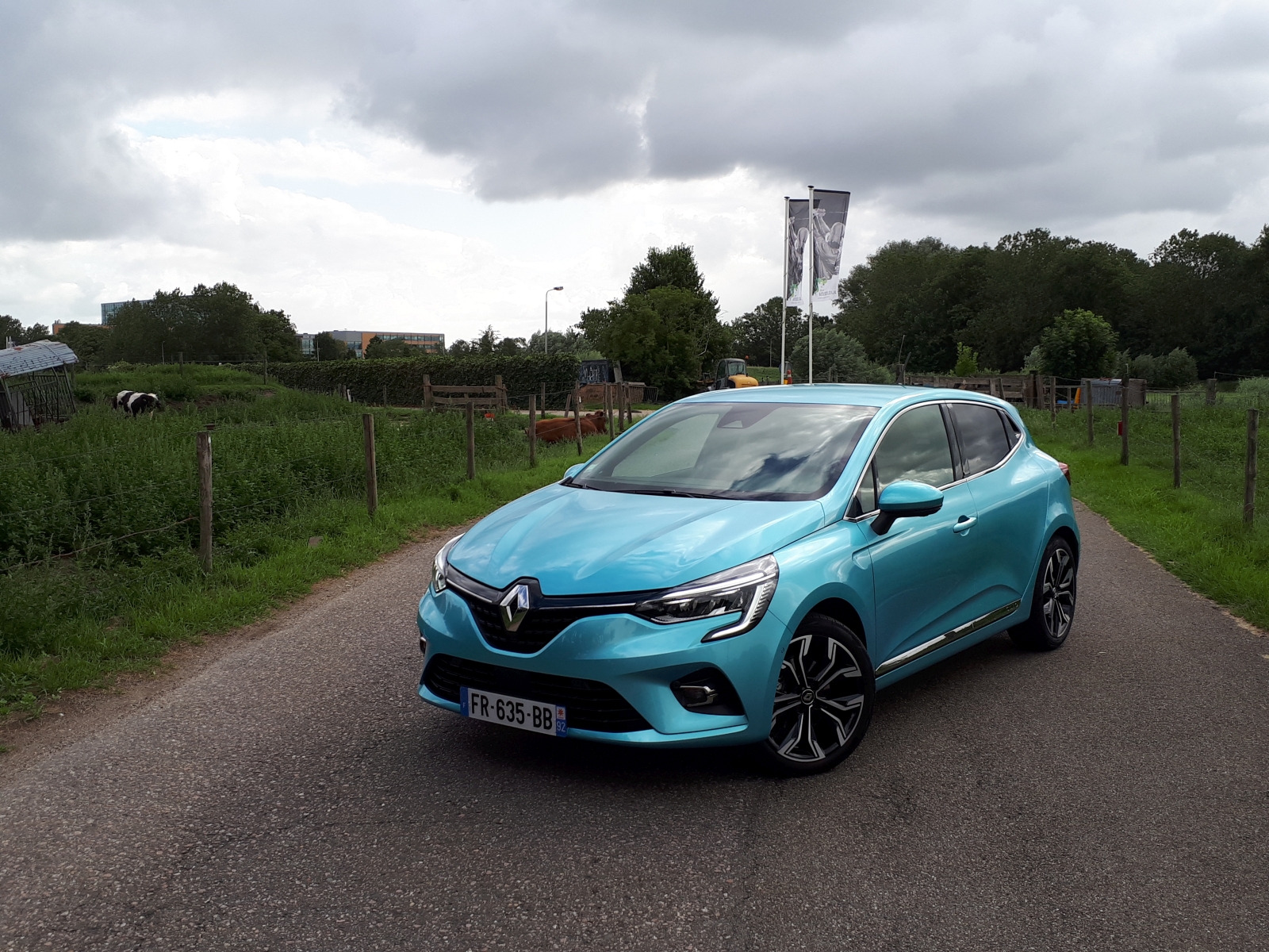 Een groenblauw Renault Clio E-Tech Hybrid geparkeerd op een landelijke weg met groene velden en bewolkte lucht op de achtergrond.