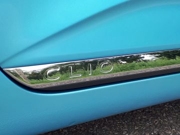 Close-up van het 'Renault Clio E-Tech Hybrid'-embleem op een turquoise autodeur, als gevolg van bomen en lucht.