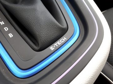 Close-up van het schakelgebied van een Renault Clio E-Tech Hybrid met het opschrift "e-tech", omgeven door een blauw verlichte rand in een zwart interieur.