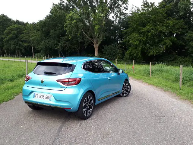 Een lichtblauwe Renault Clio E-Tech Hybrid geparkeerd langs de weg naast een grasveld met bomen op de achtergrond.