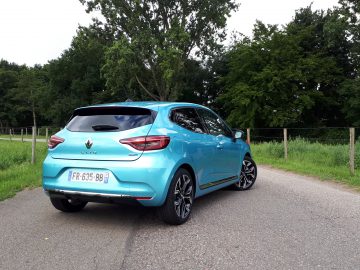 Een lichtblauwe Renault Clio E-Tech Hybrid geparkeerd langs de weg met gras en bomen op de achtergrond.