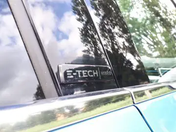 Close-up van het 'E-Tech Hybrid'-embleem van een Renault Clio op het zijpaneel van het voertuig, dat bomen en lucht weerspiegelt.