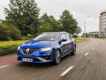 Een blauwe Renault Mégane E-Tech die op een bewolkte dag over een stedelijke weg rijdt met gebouwen op de achtergrond.