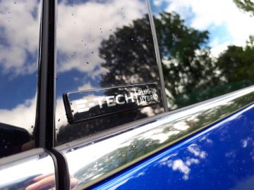 Een close-up van een 'Mégane E-Tech plug-in hybride'-badge op een blauwe auto, gezien door een reflectie op een glanzende, gedeeltelijk bewolkte dag.