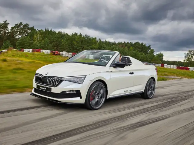 Een witte cabriolet Skoda Slavia rijdt op een racecircuit omgeven door groen gebladerte onder een bewolkte hemel.