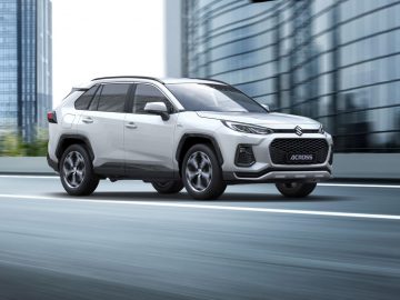 Een witte Suzuki Across SUV rijdt snel over een stadsweg met wolkenkrabbers op de achtergrond.