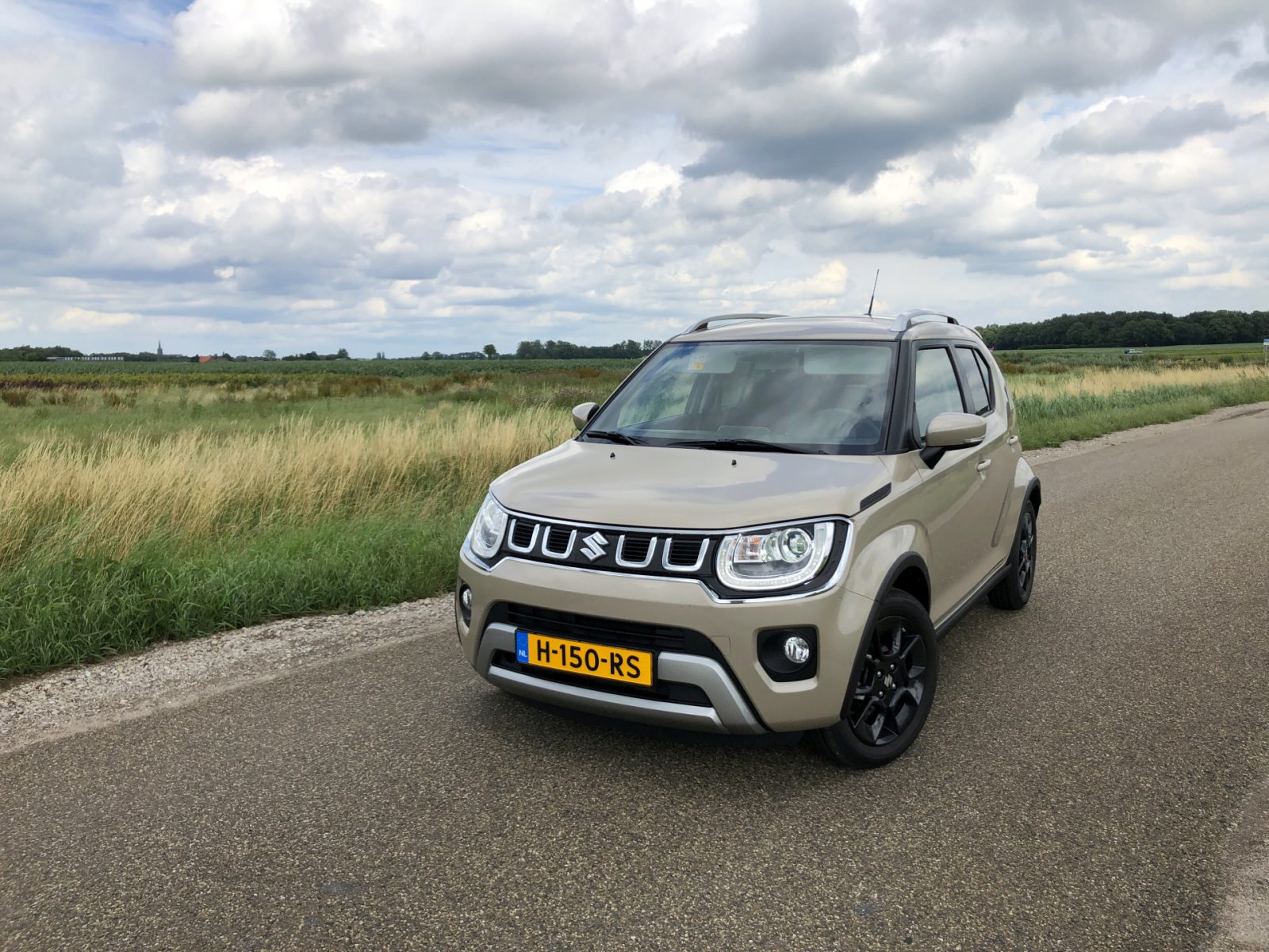 Een beige Suzuki Ignis Smart Hybrid geparkeerd op een plattelandsweg naast een veld onder een bewolkte hemel.