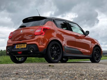 Een felrode Suzuki Swift Sport, buiten geparkeerd onder een bewolkte hemel, vanuit een lage hoek bekeken, waarbij het achter- en zijprofiel goed zichtbaar is.