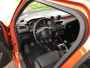 Weergave van het interieur van een Suzuki Swift Sport door een open oranje deur, met het stuur, het dashboard en de handgeschakelde versnellingsbak.