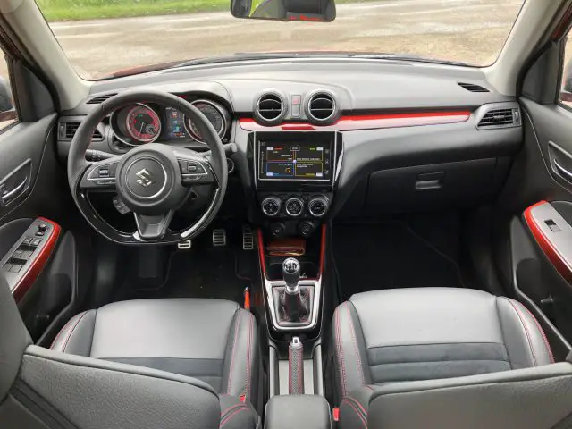 Interieur van een Suzuki Swift Sport met een stuur met logo, digitaal dashboard, middenconsole met versnellingspook en zwartleren stoelen met rode accenten.