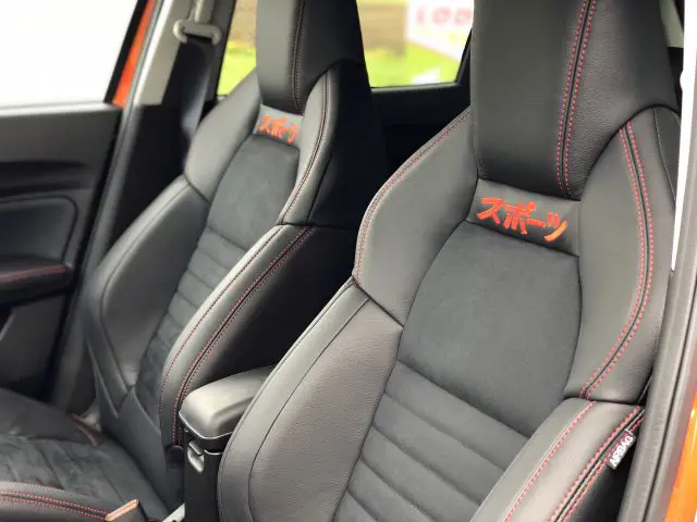 Binnenaanzicht van een Suzuki Swift Sport met twee sportieve zwartleren stoelen met oranje stiksels en logo's.