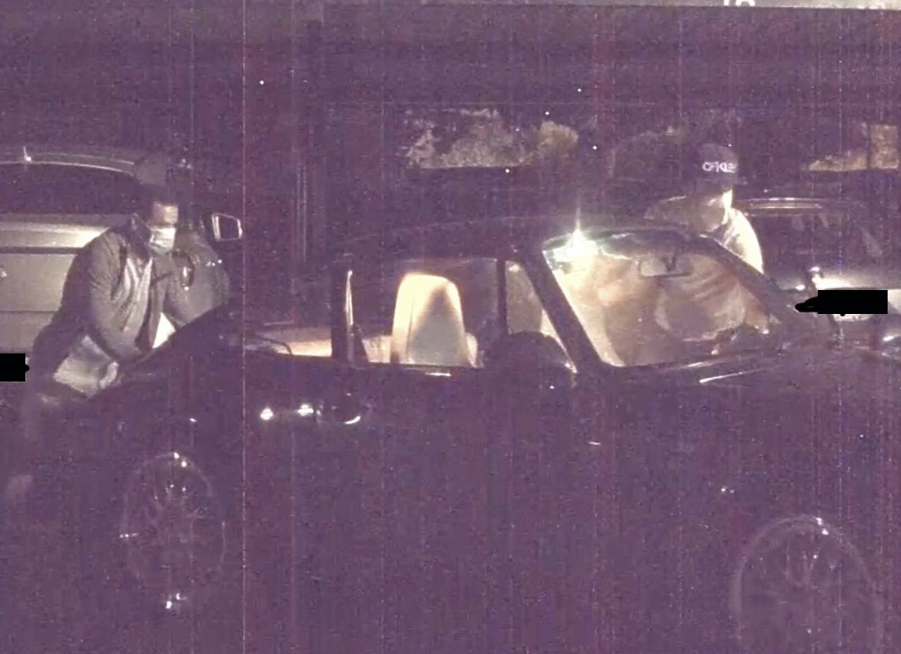 Een persoon met een masker spreekt 's nachts met een chauffeur; verlichte auto's en een donkere achtergrond op de parkeerplaats, vermoedelijk een auto gestolen.