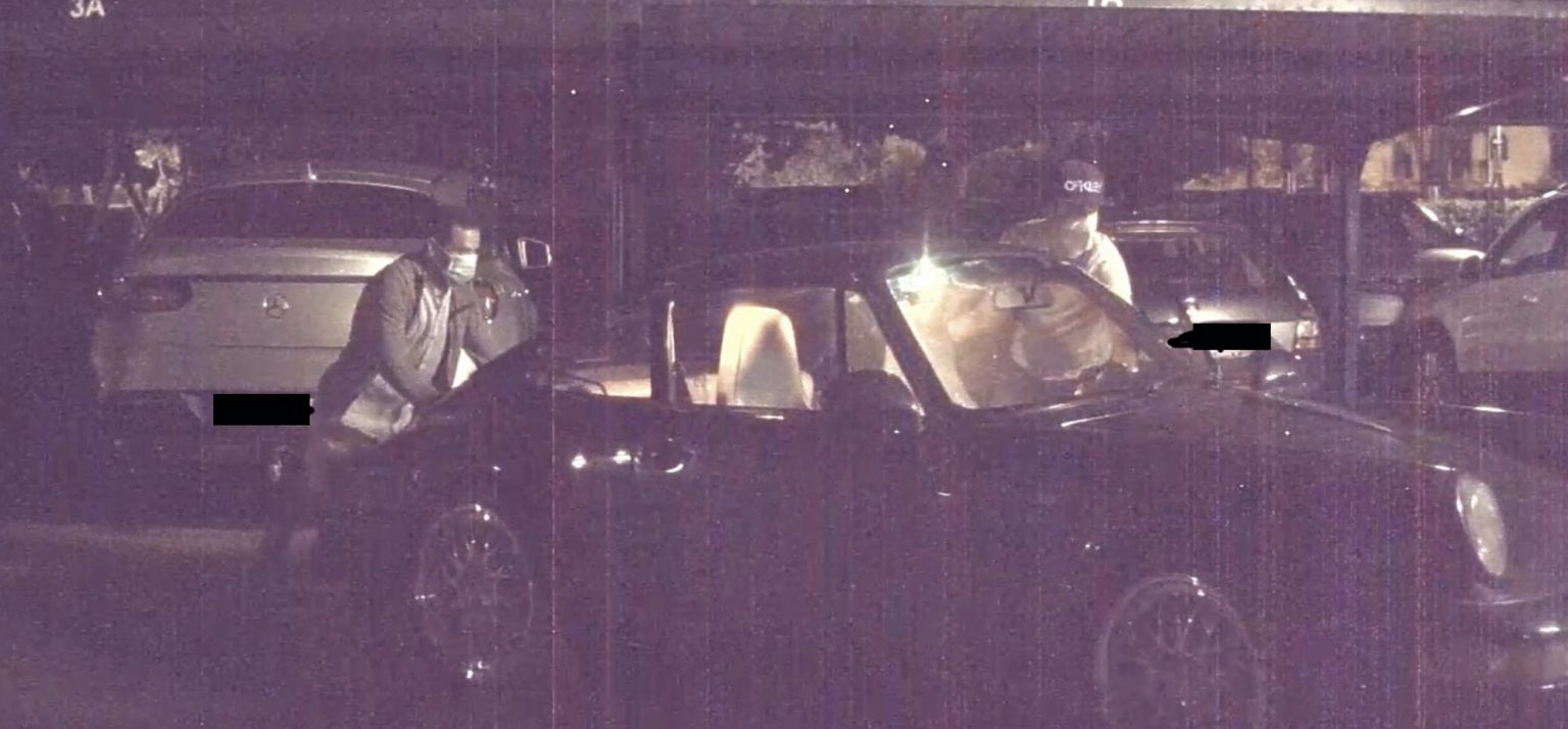 Een persoon met een masker spreekt 's nachts met een chauffeur; verlichte auto's en een donkere achtergrond op de parkeerplaats, vermoedelijk een auto gestolen.