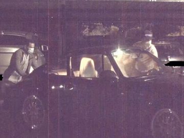 Een persoon met een masker spreekt 's nachts met een chauffeur; verlichte auto's en een donkere achtergrond op de parkeerplaats, vermoedelijk een auto gestolen.