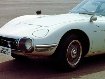 Close-up van een witte Toyota 2000GT, gefocust op de linkervoorkant, waarbij het strakke ontwerp en de uitklapbare koplampen goed te zien zijn.