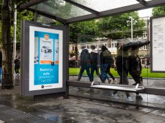 OV-reizigers lopen op een regenachtige dag langs een bushokje met digitale reclame.