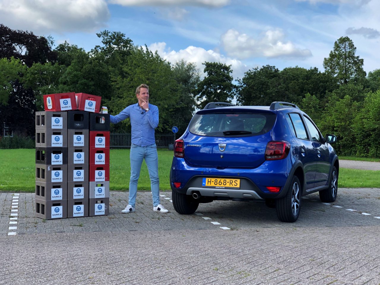 Een man die naast een stapel merkdozen stond met zijn arm omhoog, parkeerde de Dacia Sandero op de achtergrond, buiten.