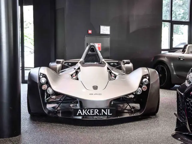 Een zilveren Ariel Atom-sportwagen tentoongesteld in een showroom met donkere vloeren en andere luxe auto's zichtbaar op de achtergrond.