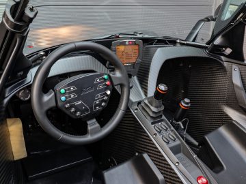 Binnenaanzicht van een Ariel Atom-sportwagen, met een stuur met bedieningselementen, een digitaal dashboard en koolstofvezelaccenten.