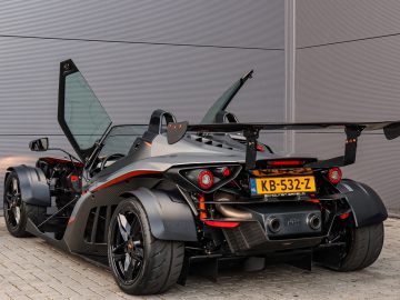 Achteraanzicht van een zwarte Ariel Atom-sportwagen met open deuren, geparkeerd tegen een grijze, gestructureerde muur.